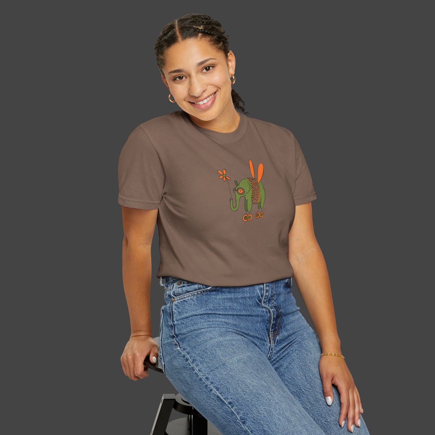 A smiling woman with braided hair sits on a stool, wearing the Flying Elephant Unisex T-shirt by Jarrod Eastman for Printify and blue jeans, against a plain dark gray background.