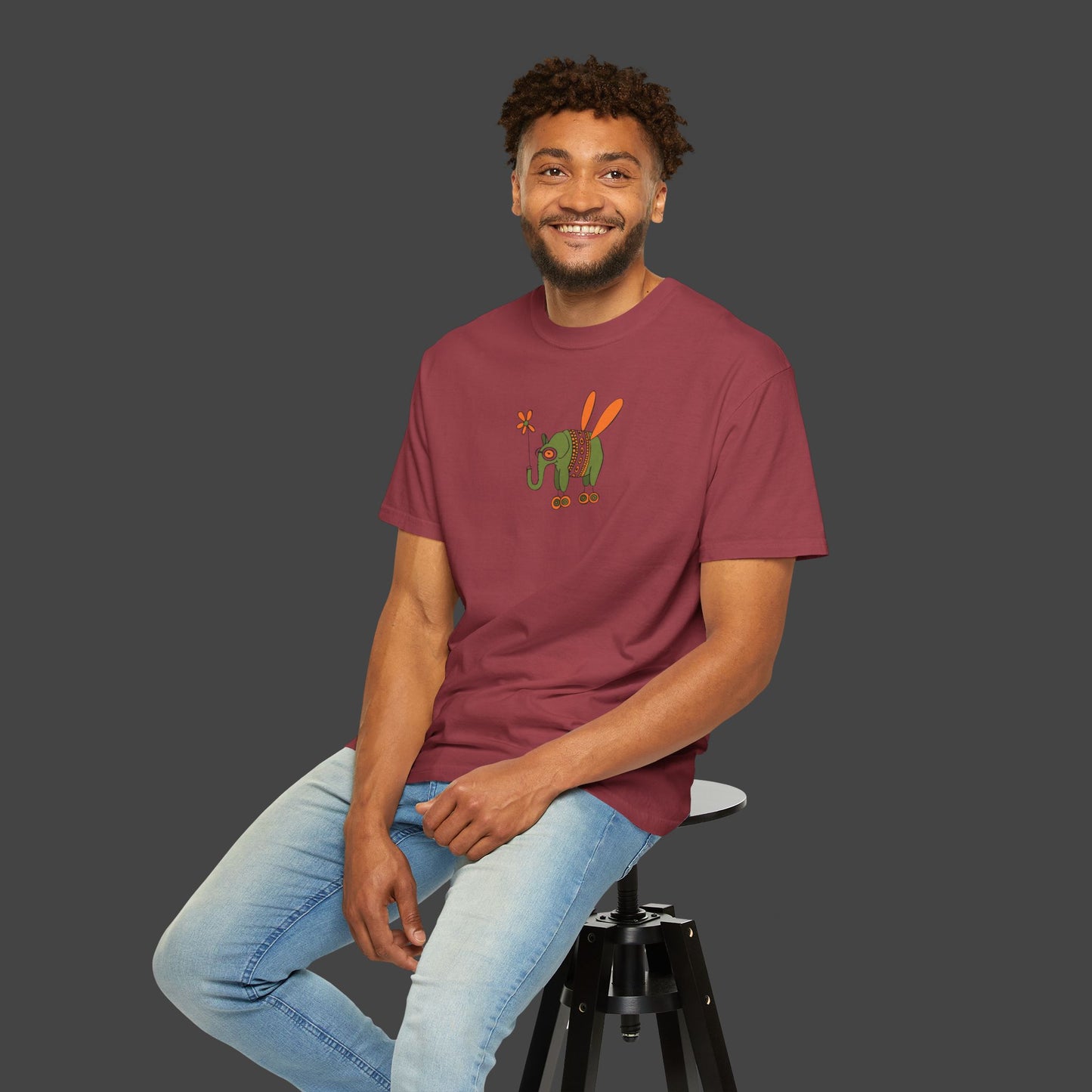 Man wearing a maroon t-shirt with a graphic design, sitting on a stool against a plain background wearing a t-shirt by the artist jarrod Eastman