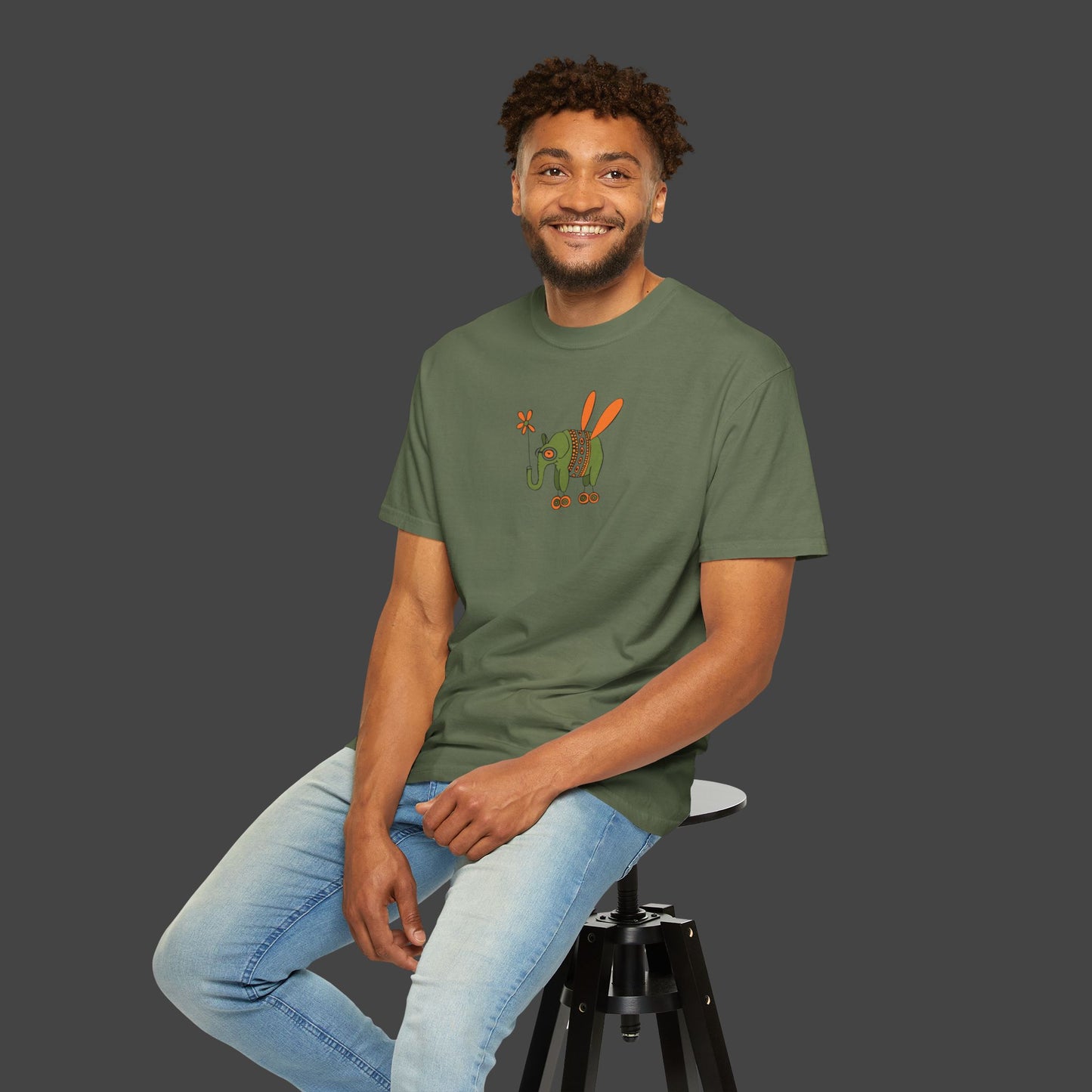 A smiling man with curly hair sits on a stool, wearing the Flying Elephant Unisex T-shirt by Jarrod Eastman for Printify and light blue jeans, set against a plain dark gray background.