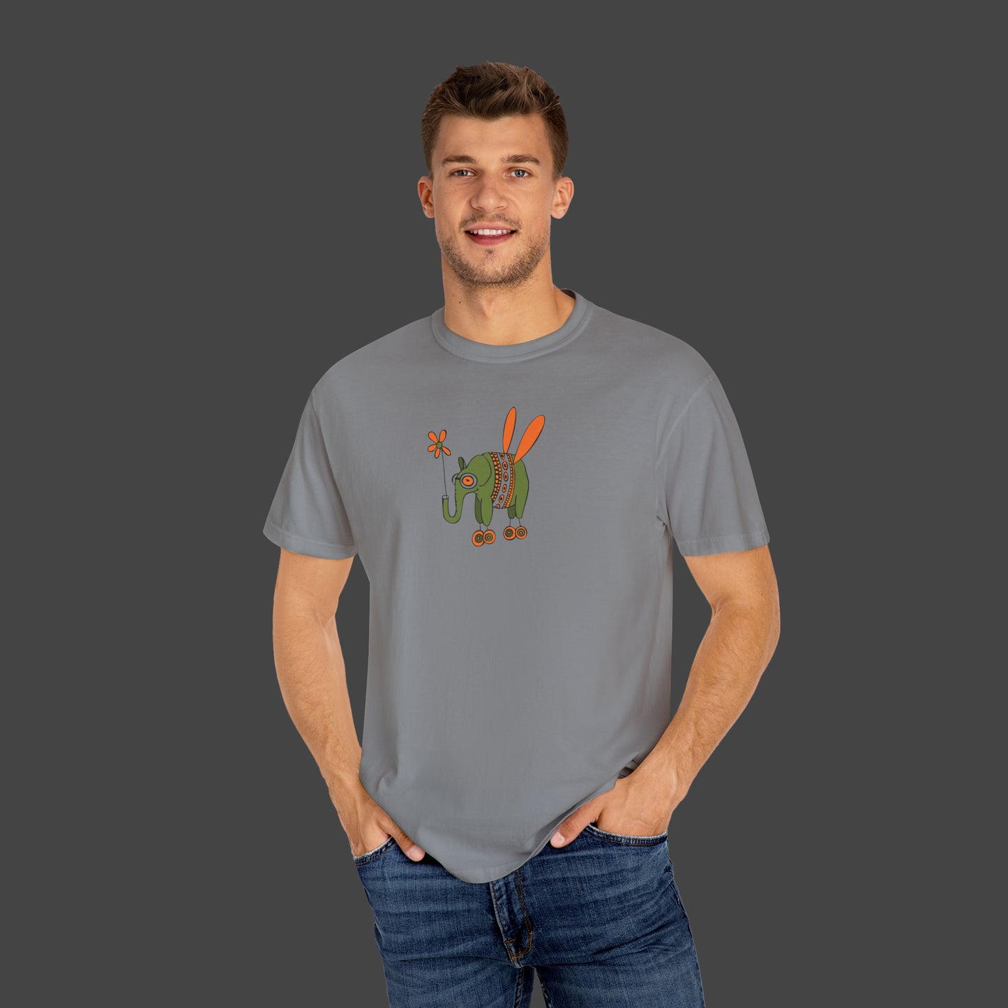 A young man wears the Printify Flying Elephant Unisex T-shirt by Jarrod Eastman, featuring a colorful cartoon elephant with orange wings and wheels, smiling against a dark gray background.