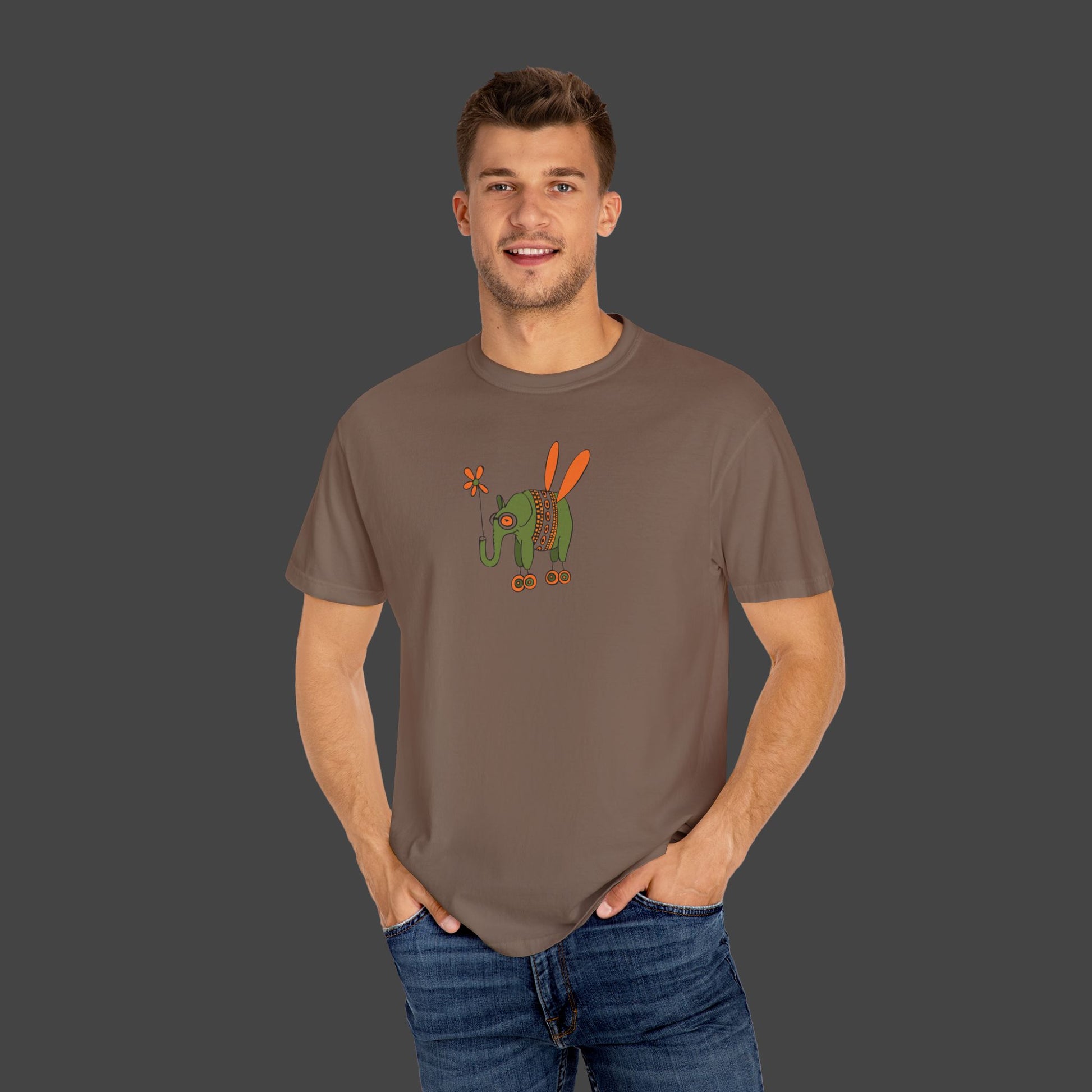 A smiling man wears the Flying Elephant Unisex T-shirt by Printify, designed by Jarrod Eastman, featuring a whimsical green elephant with orange wings and a flower, set against a plain gray background.