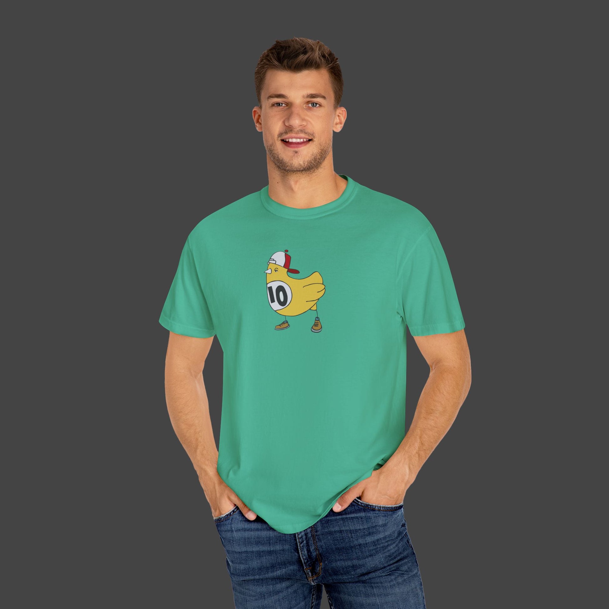 A smiling man wears the Chicken Scratch Unisex T-shirt by Jarrod Eastman (Printify)—a teal, garment-dyed tee with quirky art of a helmeted duck and number 10—standing hands-in-pockets against a plain gray background.