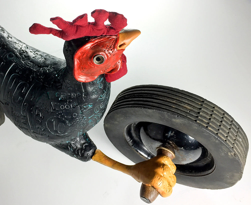 Black Chicken by jarrodeastman is a whimsical sculpture featuring a dark, patterned rooster with a red comb, holding a large black tire in one wing. Crafted from found objects, it stands out against a plain white background.