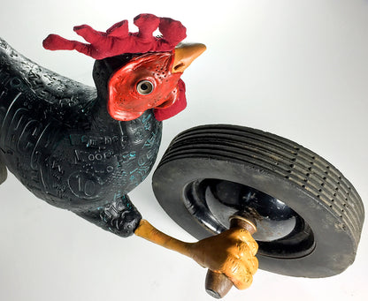 Black Chicken by jarrodeastman is a whimsical sculpture featuring a dark, patterned rooster with a red comb, holding a large black tire in one wing. Crafted from found objects, it stands out against a plain white background.