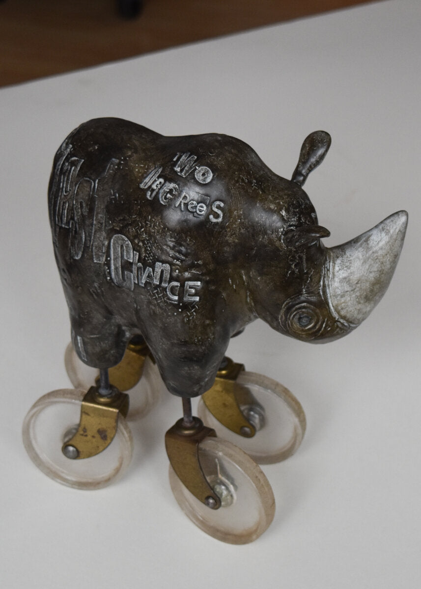 The Heavy Weight 6/30 by jarrodeastman is a bronze rhinoceros sculpture on four caster wheels, each with metal brackets, engraved with detailed letters for a rolling, whimsical effect.