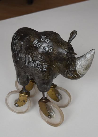 The Heavy Weight 6/30 by jarrodeastman is a bronze rhinoceros sculpture on four caster wheels, each with metal brackets, engraved with detailed letters for a rolling, whimsical effect.