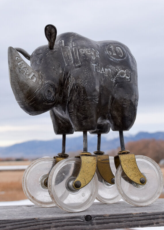 Heavy Weight 6/30 by jarrodeastman is a whimsical bronze rhinoceros sculpture set on four found castor wheels, with engraved words on its side and mountains in the background under a cloudy sky.