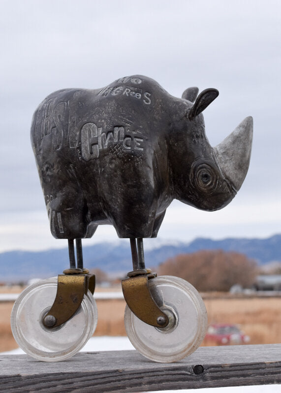 Heavy Weight 6/30 by jarrodeastman is a metal sculpture of a rhino with two large found castor wheels as legs, standing on a wooden rail outdoors with mountains and a cloudy sky in the background.