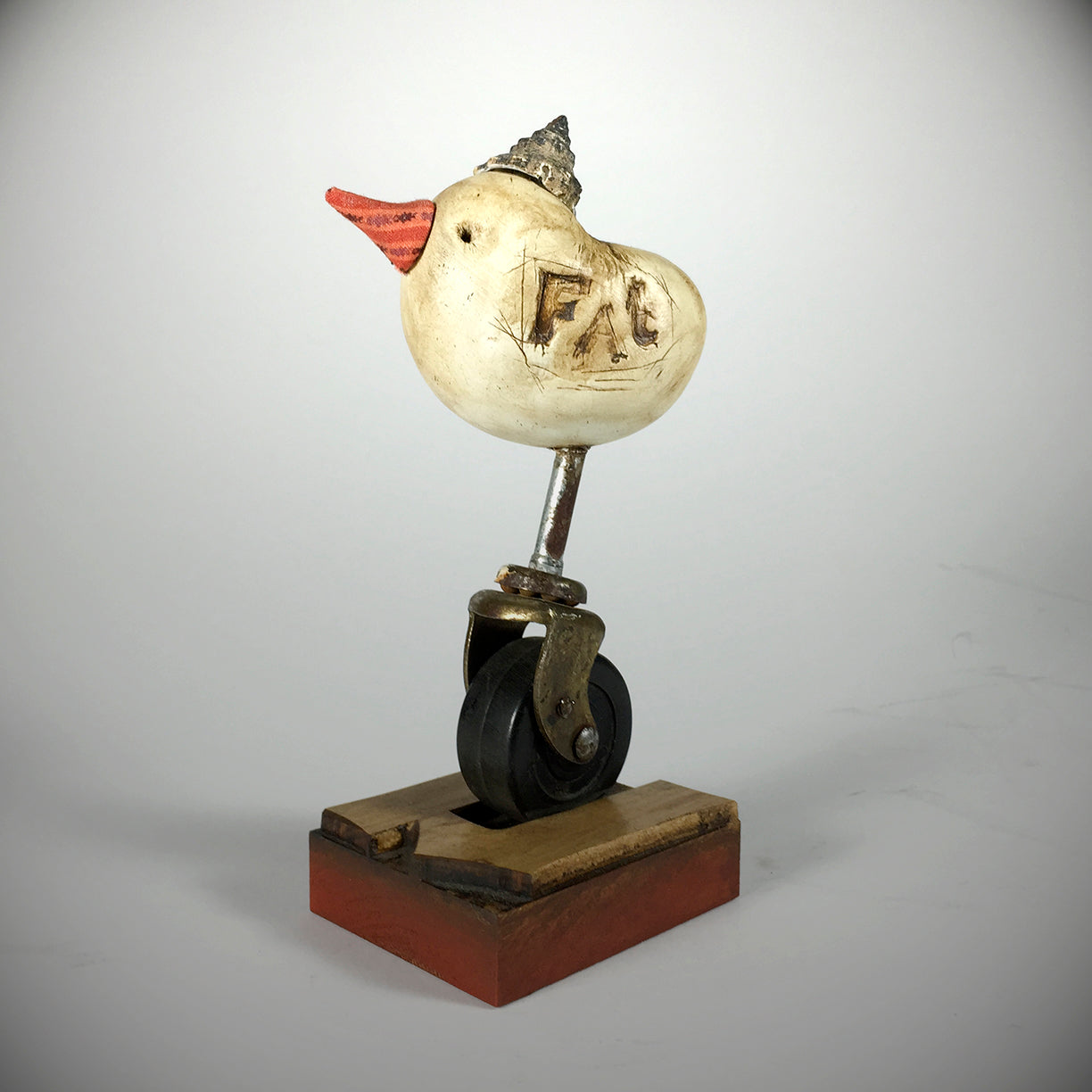 Fat by jarrodeastman is a small original polymer clay sculpture of a duck with an orange beak, F.A.B. carved on its side, a tiny metallic cone hat, perched on a metal caster wheel and mounted on a wooden base.