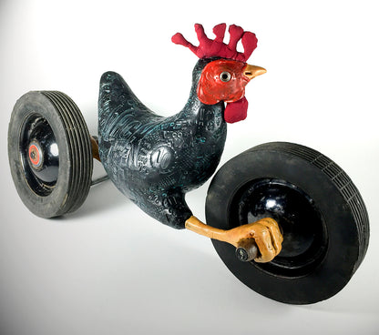 The jarrodeastman Black Chicken is a whimsical sculpture featuring a black chicken with a red comb and yellow beak, mounted on two large antique wheels for a playful, cart-like look.