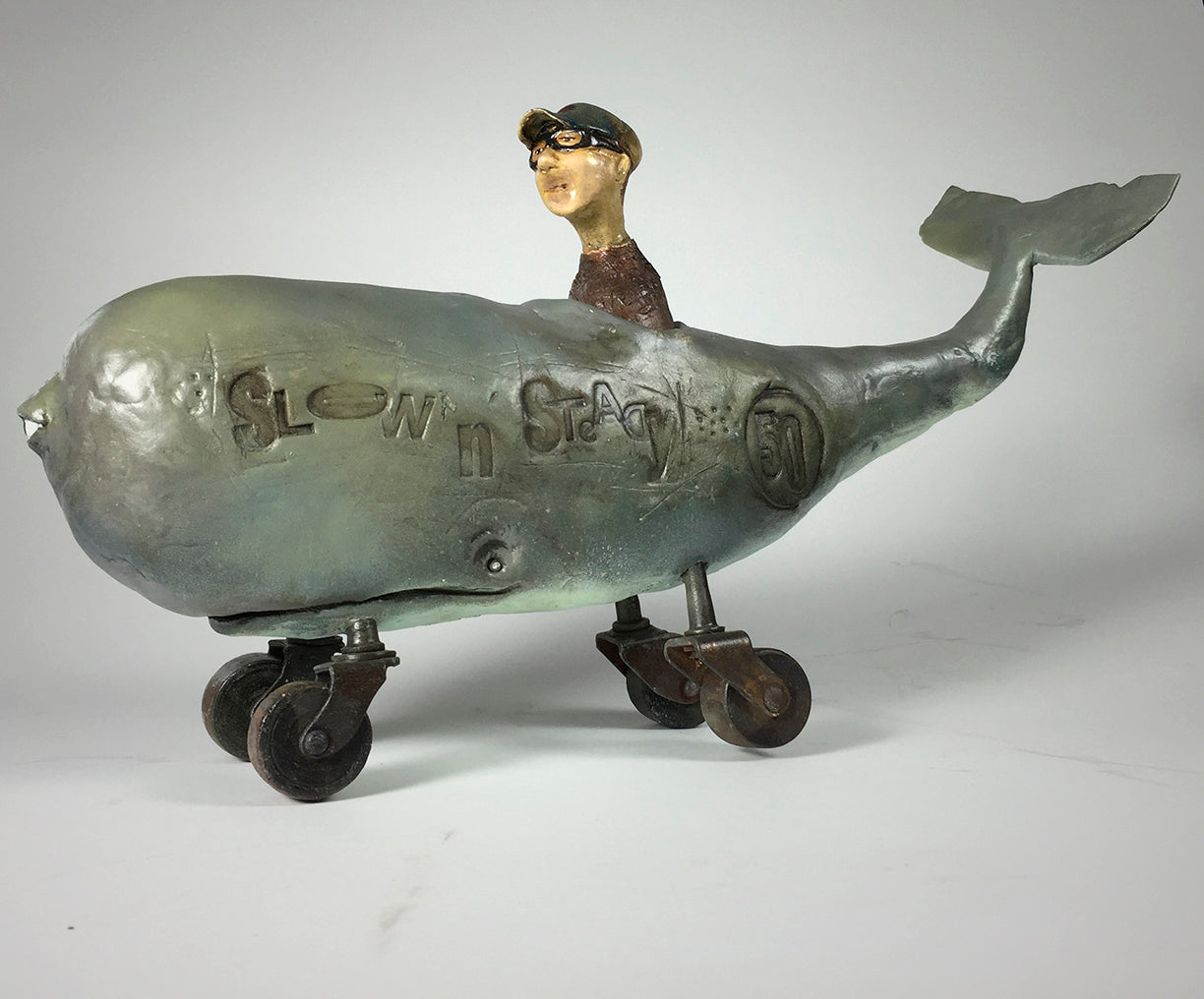 Derby by jarrodeastman: A whimsical polymer clay sculpture of a whale on antique castor wheels, featuring a glasses-wearing, capped figure emerging from its back; SLOW n steady 50 inscribed on the side.