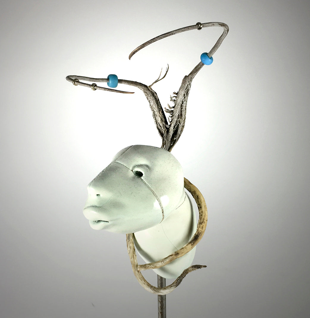 The Earth sculpture by jarrodeastman is a clay animal head with a smooth, pale finish, slim antlers decorated with blue beads, and a curved horn-like form wrapping from the neck. Set against a plain, grayish-white background.
