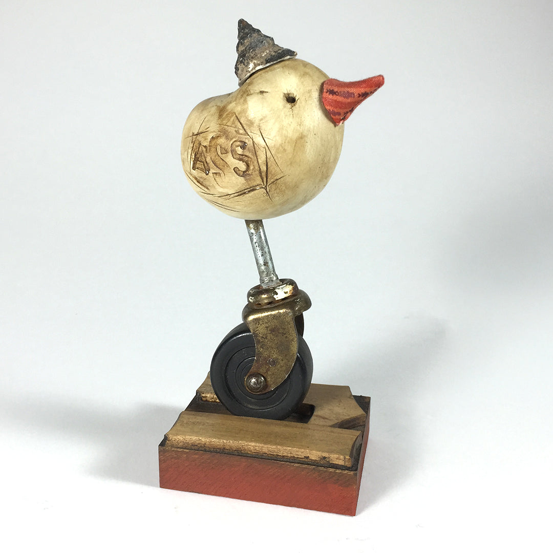 Fat by jarrodeastman is a small whimsical polymer clay sculpture of a round-bodied bird with an orange beak, gray hat, and engraved letters. It sits on a caster wheel mounted to a wooden base.