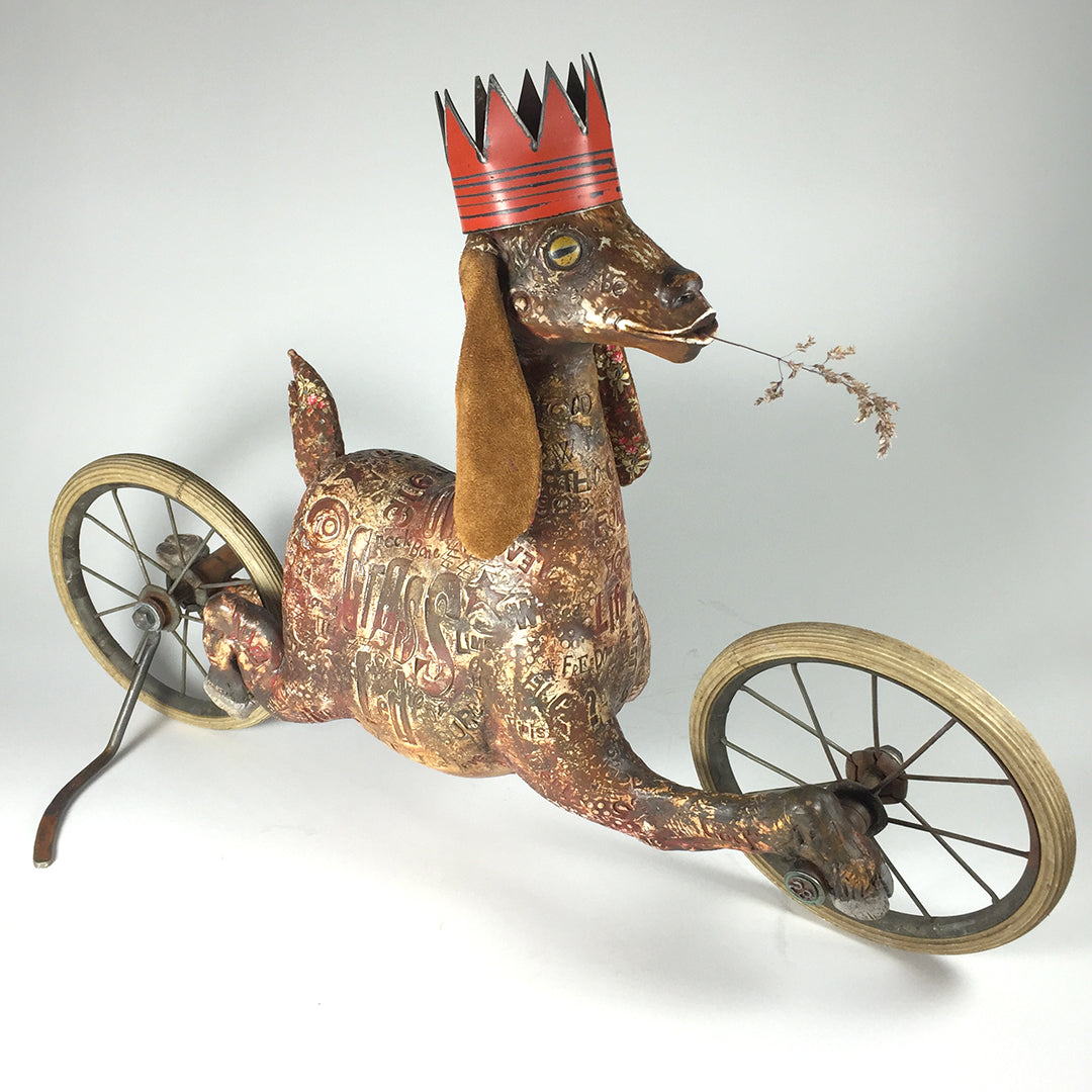 A whimsical, vintage-style original sculpture by jarrodeastman, Goat features antique wheels, etched words and patterns, a red crown, and a twig in its mouth—evoking the nostalgic charm of a rolling toy.
