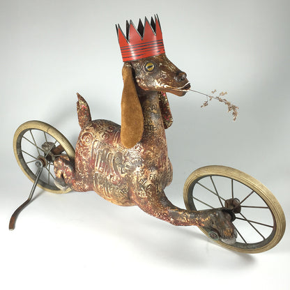A whimsical, vintage-style original sculpture by jarrodeastman, Goat features antique wheels, etched words and patterns, a red crown, and a twig in its mouth—evoking the nostalgic charm of a rolling toy.