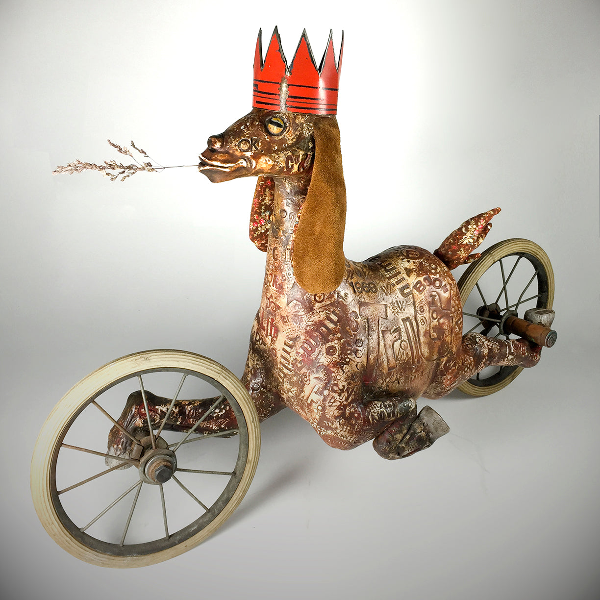 Goat by jarrodeastman is a whimsical, dog-like metal sculpture with circuit patterns, a red crown, brown felt ears, a twig in its mouth, and mounted on three antique wheels.