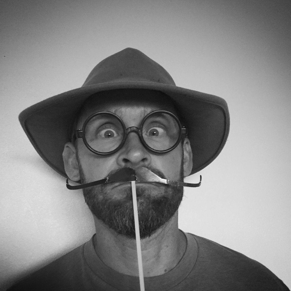 A man wearing a wide-brimmed hat and round glasses poses with a fake mustache on a stick, making a comical, wide-eyed expression against a plain background.  That is Jarrod Eastman!