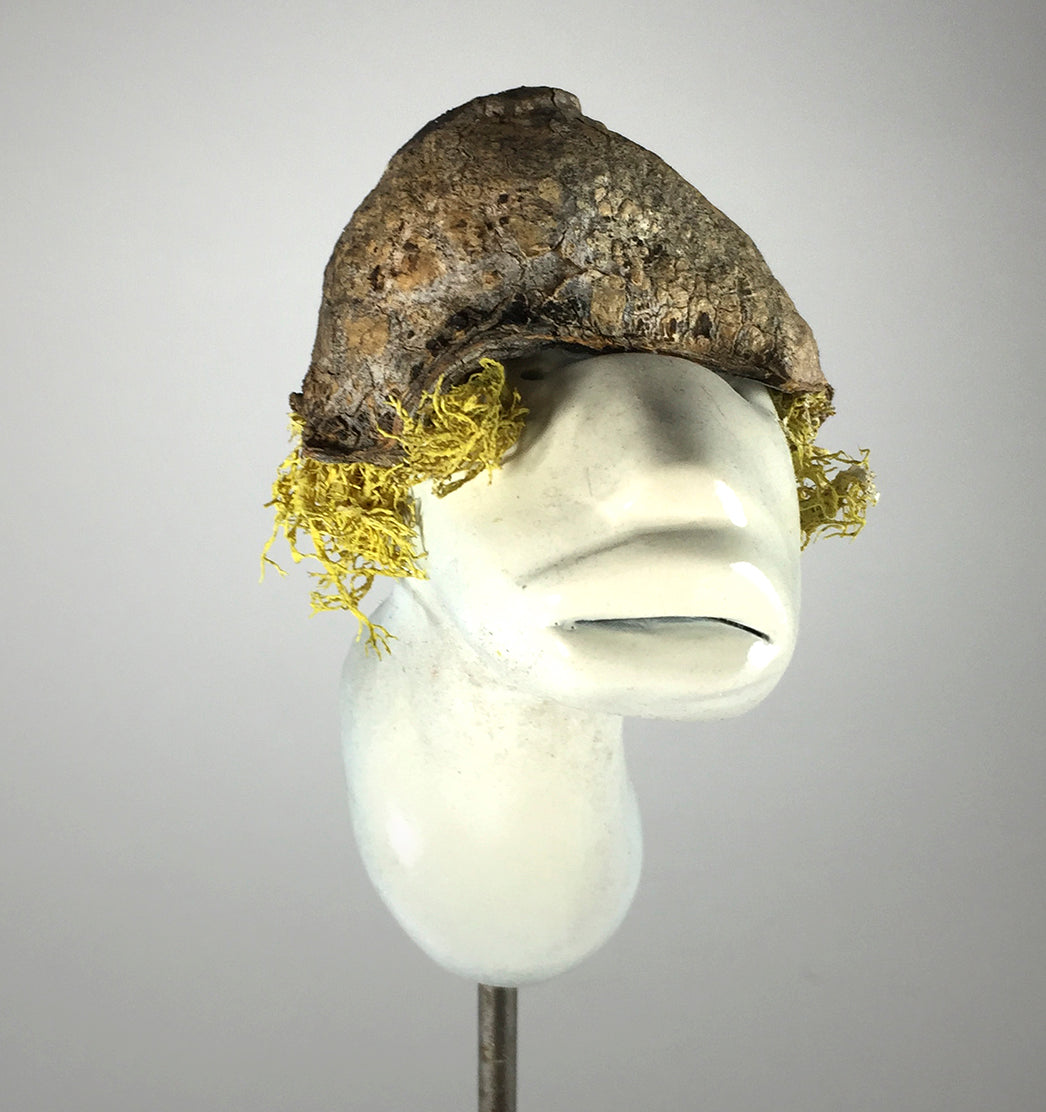 A white mannequin head displays Seed by jarrodeastman—a hat sculpted from rough brown tree bark with yellow moss hair. Set against a plain light gray background.