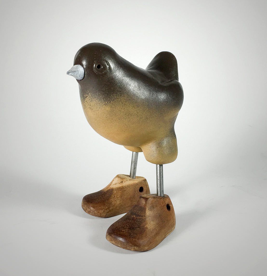 The Shoes sculpture by jarrodeastman is a small bird with a smooth, dark body and wooden legs adorned with charming shoes, set against a plain light background.
