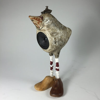 The Tweeter by jarrodeastman is a whimsical bird sculpture speaker with a patterned beak, ornate crown, long white legs wrapped in red thread, and mismatched wooden shoes, set against a plain background.