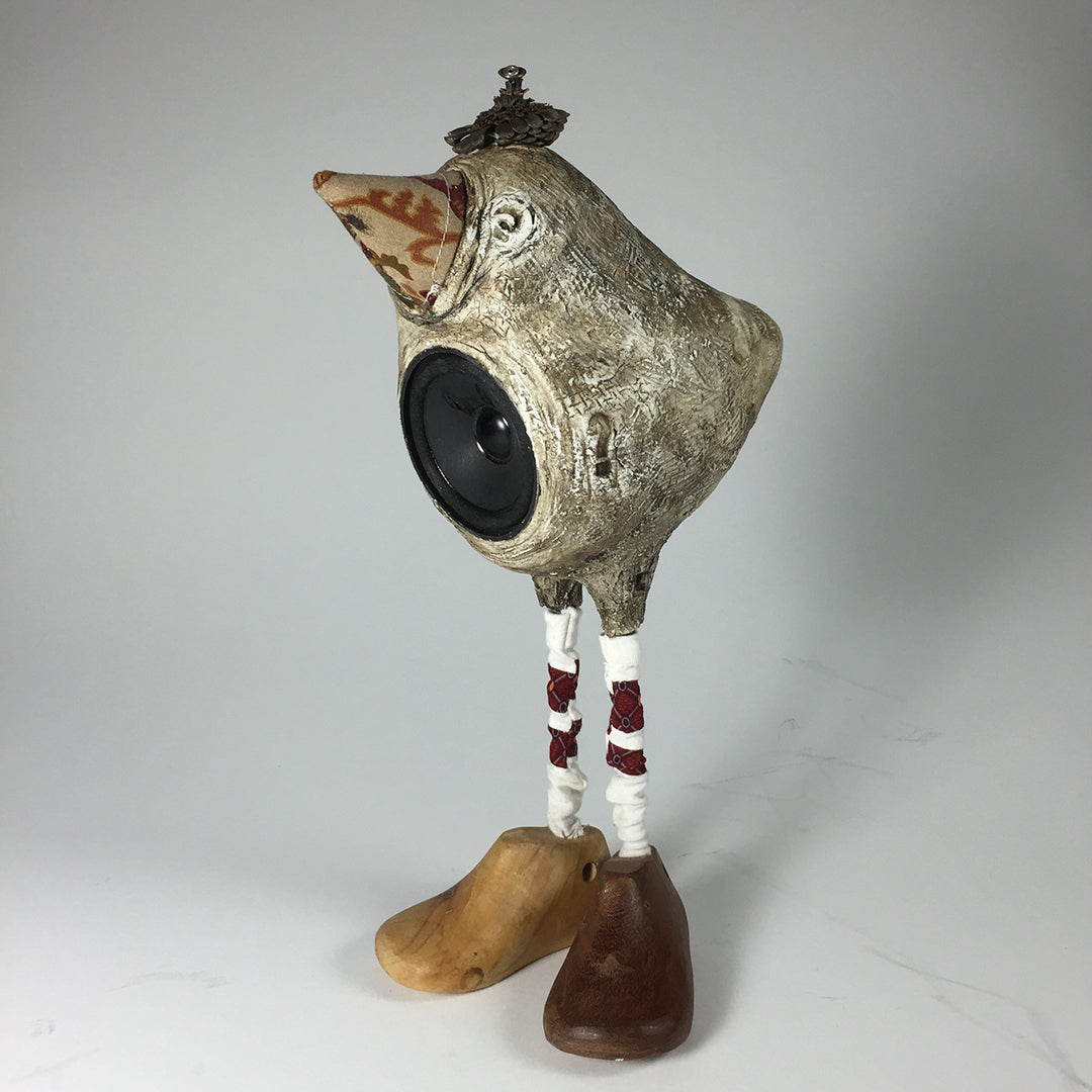 The Tweeter by jarrodeastman is a whimsical bird sculpture speaker with a patterned beak, ornate crown, long white legs wrapped in red thread, and mismatched wooden shoes, set against a plain background.