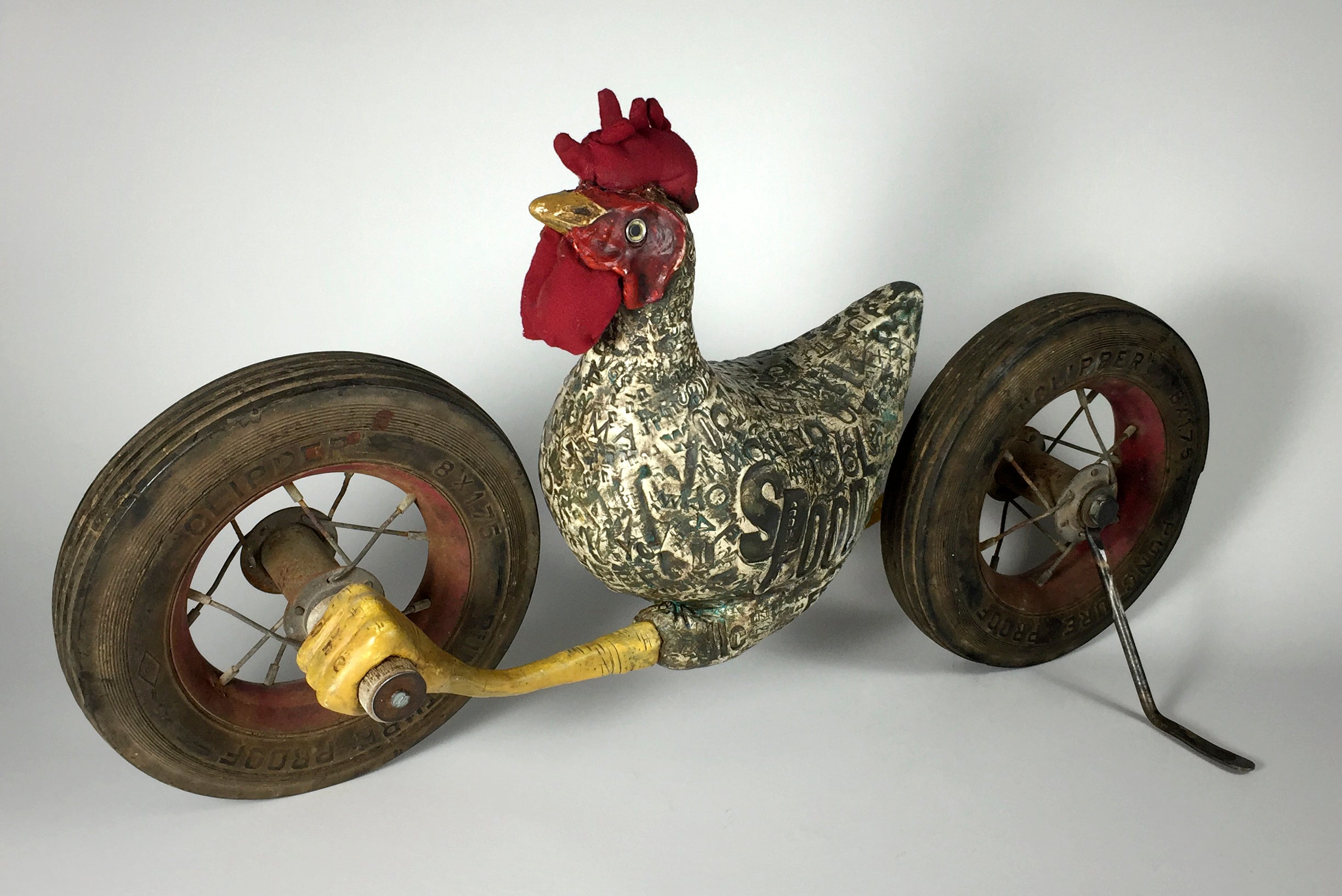 The jarrodeastman White chicken is a whimsical sculpture featuring a patterned body, red fabric comb, and two large antique wheels, resembling a quirky tricycle against a plain background.