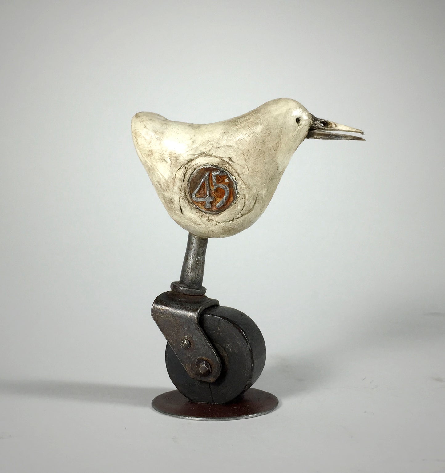 Why by jarrodeastman: a small original polymer clay sculpture of a white bird with an orange 45 on its side, standing on an antique castor wheel, displayed against a plain gray background.